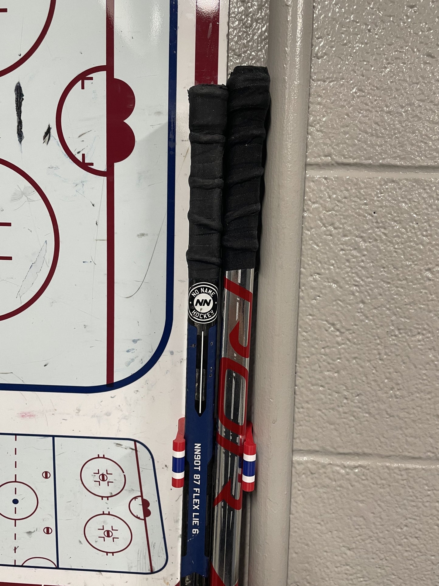 Stick Em Up Hockey Stick Clip Holding Two Sticks Aligned in Front of a Locker Room's Practice Board